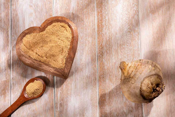 Maca-Pulver auf einem Holzlöffel und in einer herzförmige Holzschale sowie eine Maca-Wurzel auf einem Holztisch.