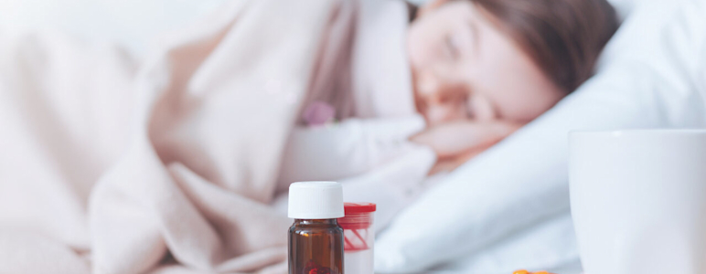 Schlaftabletten stehen auf einem Nachttisch, im Hintergrund schläft eine Frau im Bett.