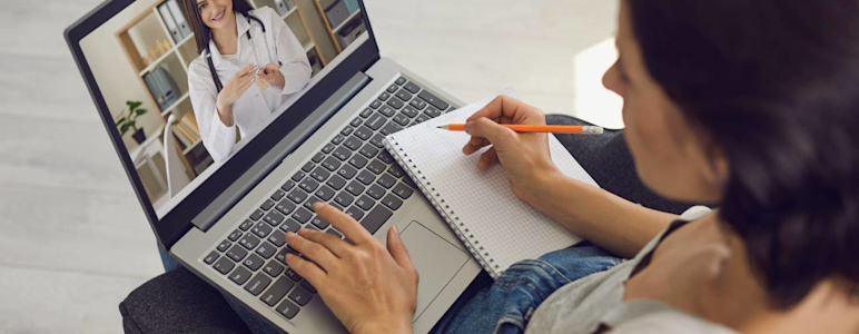 Eine Frau sitzt am Laptop und macht sich während der Online-Sprechstunde mit einer Ärztin Notizen.