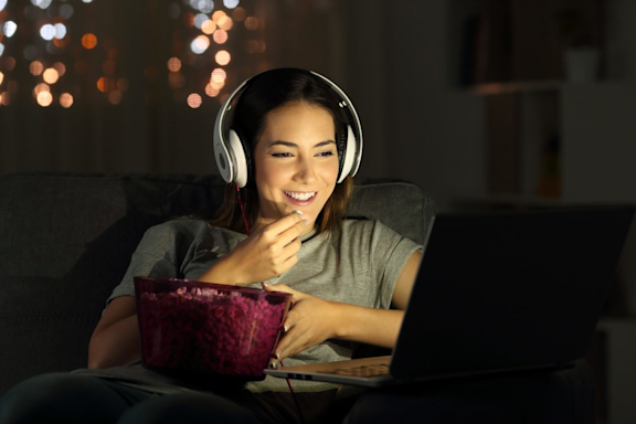 Es ist eine junge Frau zu sehen. Sie trägt Kopfhörer und isst vor ihrem Laptop Popcorn.