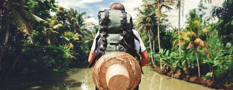 Frau macht einen Abenteuerurlaub. Sie steht mit Rucksack und Strohhut an einem Fluss.