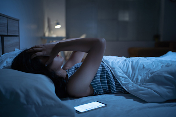 Junge Frau liegt nachts schlaflos im Bett, neben ihr ein Handy.