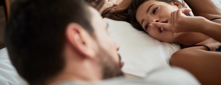 Mann und Frau liegen im Bett. Die Frau hält ihren Zeigefinger an ihre Lippen.