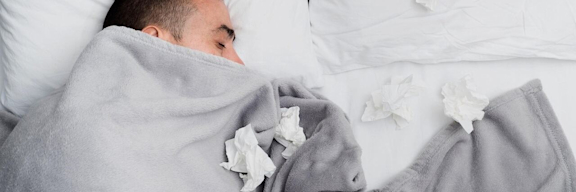 Ein Mann liegt im Bett. Er ist bis zur Nase zugedeckt und es liegen viele benutzte Taschentücher um ihn herum.