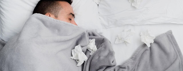 Ein Mann liegt im Bett. Er ist bis zur Nase zugedeckt und es liegen viele benutzte Taschentücher um ihn herum.