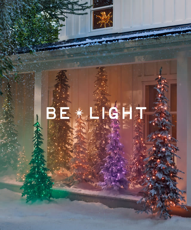 Illuminated Christmas trees in various colors displayed on a snowy porch with "BE LIGHT" text overlay and twinkling lights.