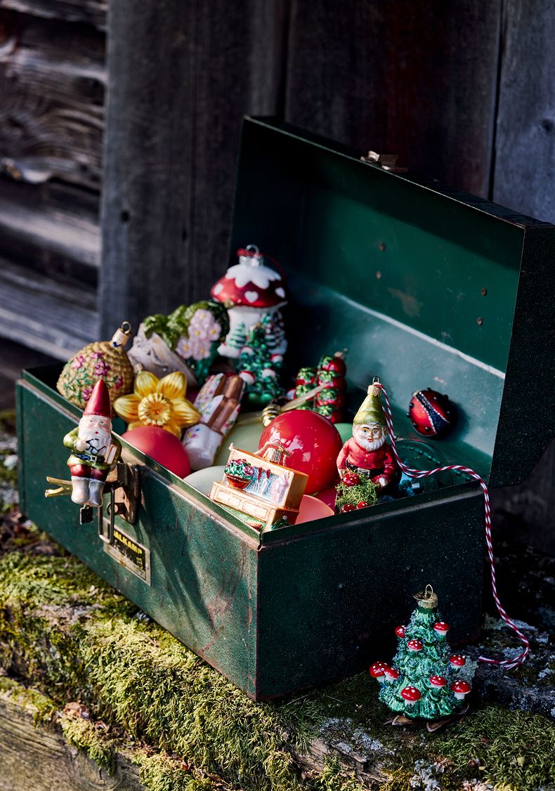 Green metal toolbox filled with vintage Christmas ornaments including Santa figures, trees, and colorful decorations on mossy ground.