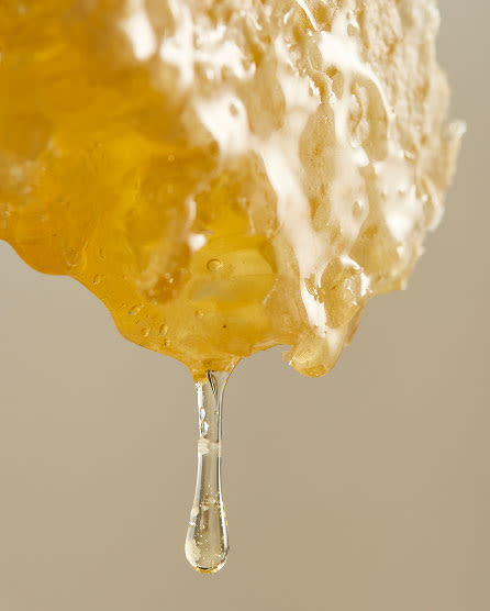 Golden honey dripping from a honeycomb piece against a neutral beige background, showing the thick, viscous texture.
