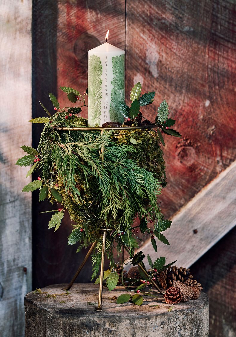 White pillar candle with green fern design sits atop evergreen wreath on gold stand with pinecones against rustic backdrop