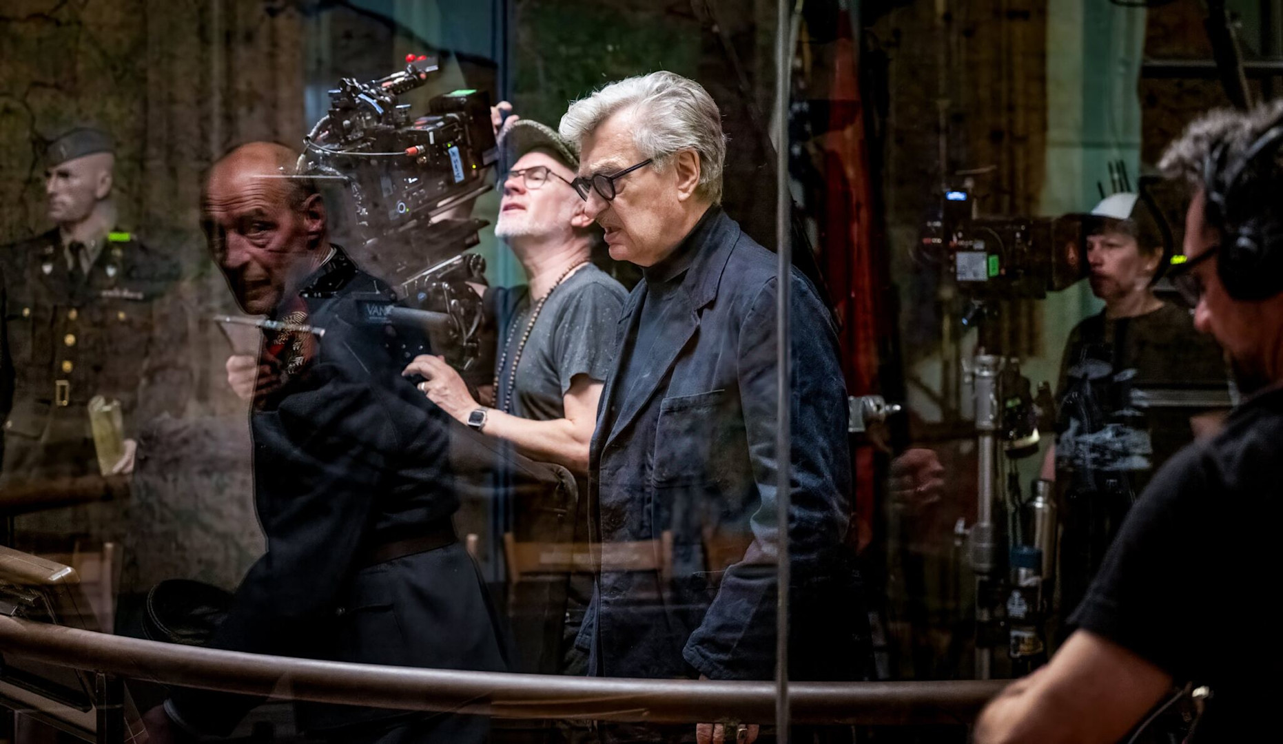 Case | Wim Wenders und die die Schlüssel zur Freiheit | Auswärtiges Amt | Image Regisseur Wim Wenders steht in dunkler Kleidung nachdenklich an einem Filmset. Um ihn herum sind ein Kameramann, weitere Crewmitglieder und eine Schaufensterpuppe in einer historischen Uniform durch eine spiegelnde Scheibe zu sehen.