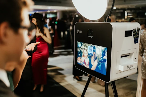 Selfie Zone szelfigép prémium háttér előtt, a képernyőjén vicces kellékekkel mosolygó vendégekkel.