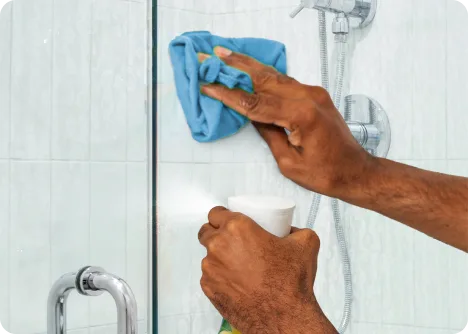 Personne nettoyant une paroi de douche en verre avec un chiffon bleu tout en tenant un vaporisateur dans l’autre main.