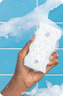 Main tenant un flacon pulvérisateur blanc à motif géométrique, recouvert de mousse de savon, sur un fond de carreaux bleus clairs.