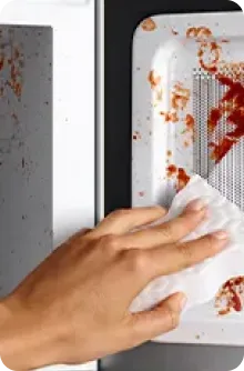 Main essuyant des éclaboussures de sauce rouge sur la paroi intérieure blanche d’un micro-ondes à l’aide d’un essuie-tout.