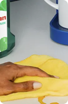 Main essuyant un comptoir avec un chiffon en microfibre jaune, à côté d’un flacon de produit nettoyant et d’un porte-essuie-tout.