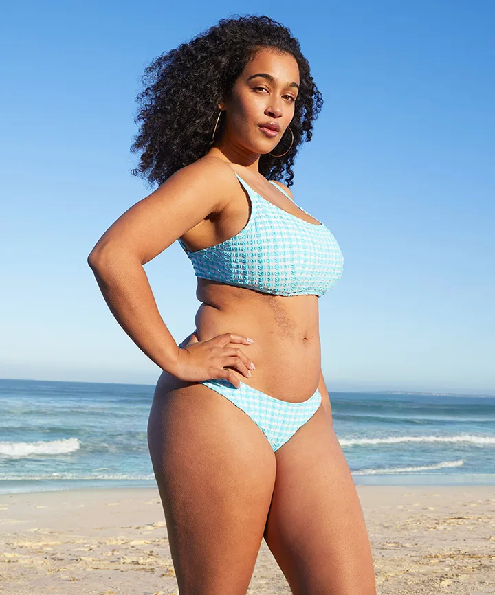 A woman with curly hair stands on the beach wearing a blue checkered bikini, with ocean waves in the background.