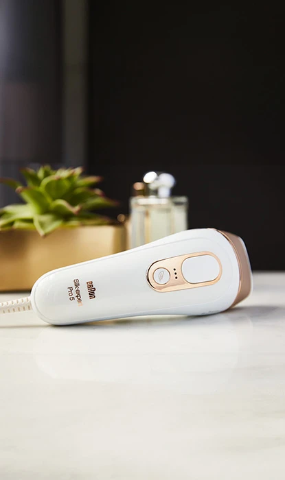 Braun IPL device with a white and gold design on a marble countertop, with plants and beauty products in the background.