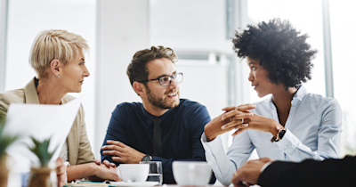 Three financial advisors are sitting at a table drinking coffee with bright windows in the background.