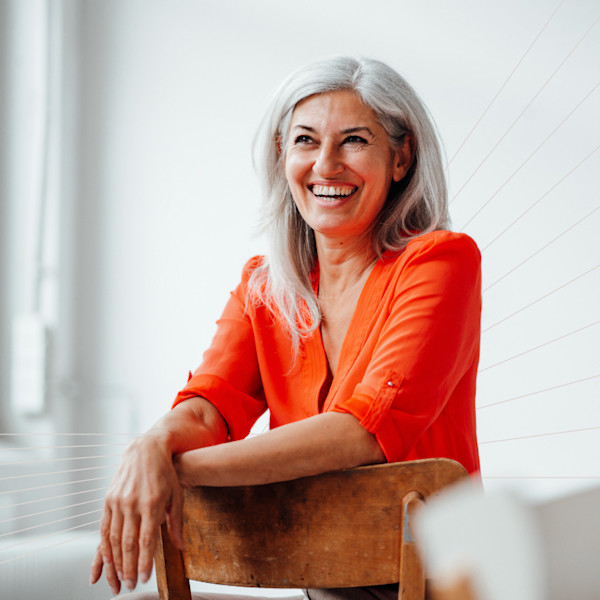 A woman wearing a tomato red shirt sits backward on a chair, smiling.