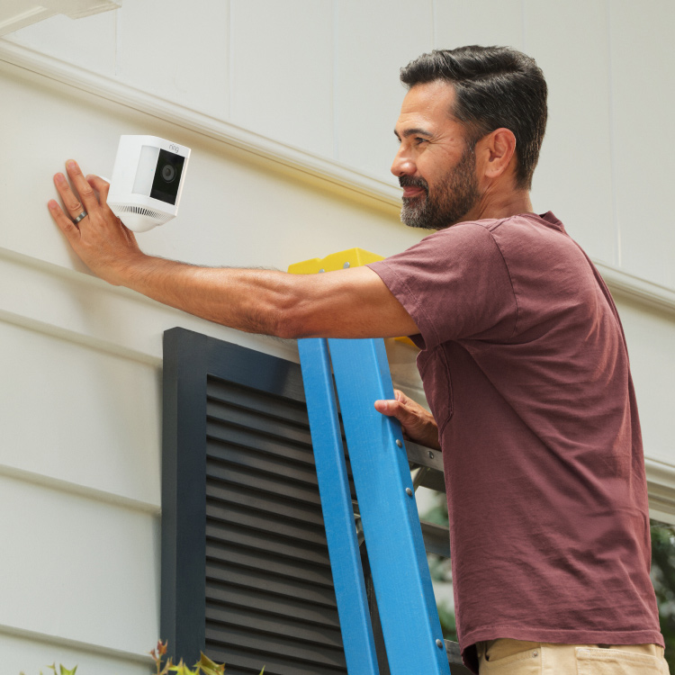 Man on a ladder mounting a white Spotlight Cam Plus on a home exterior wall using a white PoE plus Converter Mount.