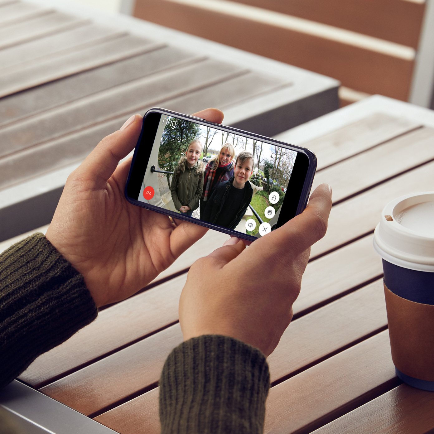 Two hands holding a mobile phone. On the phone screen is a video of three people standing in front of a closed door.
