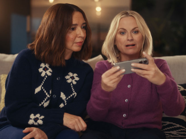 Two women sit on a couch, one in a purple sweater holding a smartphone while the other in a navy sweater watches, reacting to something on the screen.