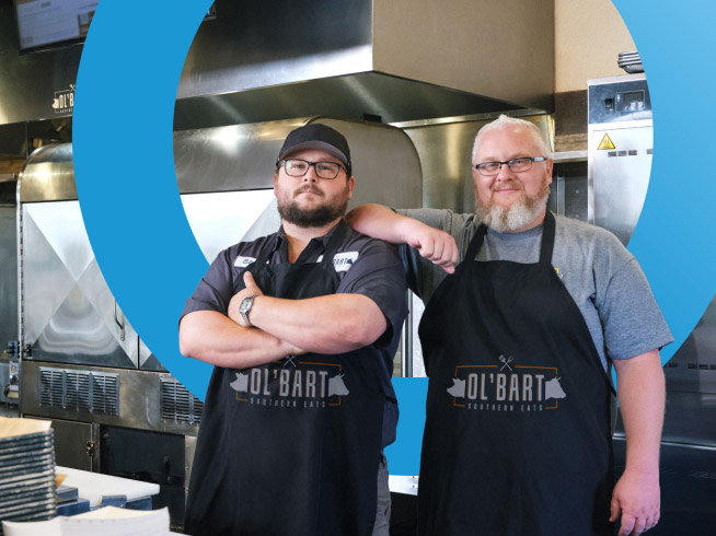 Two restaurant owners wearing Ol' Bart's Southern Eats aprons stand confidently in their kitchen.