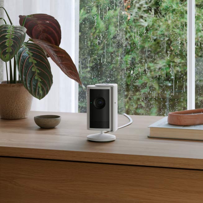 White Indoor Cam Plus sits on a surface indoors next to decorative books and a potted plant.