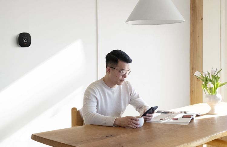 A man in a white shirt sits at a table drinking tea and looking at his mobile phone. A Smart Thermostat Premium is attached to the wall behind him.