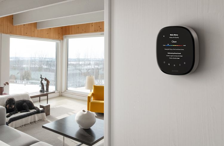 An ecobee Smart Thermostat is mounted to a white wall in the foreground. The thermostat displays a screen that indicates that the air quality is currently "clean". A room with minimalist white furniture and two large windows is visible past the wall.