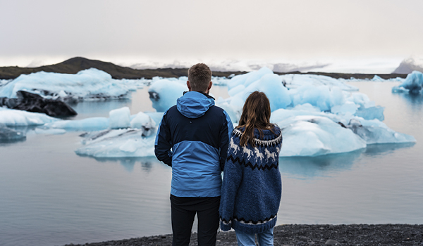 Two people, one in a blue jacket and the other in a traditional Icelandic sweater, standing together overlooking a stunning glacial lagoon with floating icebergs, symbolizing exploration and companionship.