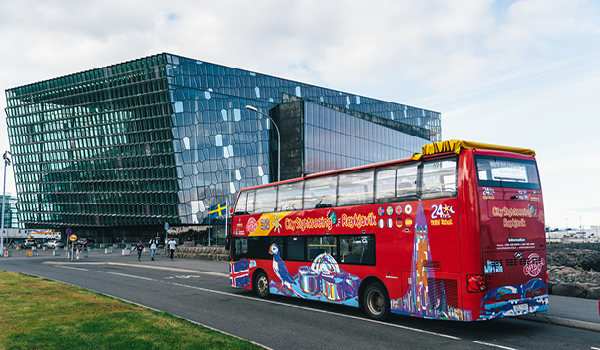 Red hop-on hop-off tourist bus in front of the Harpa Reykjavík Concert Hall on a clear day..
