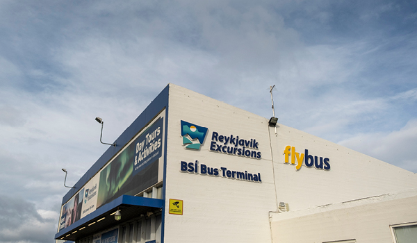 The BSI Bus Terminal building under a partly cloudy sky, adorned with signs for 'Reykjavik Excursions' and 'Flybus,' indicating it as a hub for travelers in Reykjavik