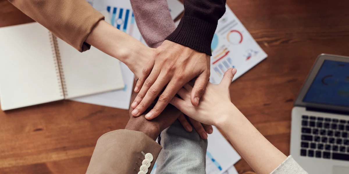 Businesspeople pile their hands in a circle over charts and notes, signifying their commitment to their team.