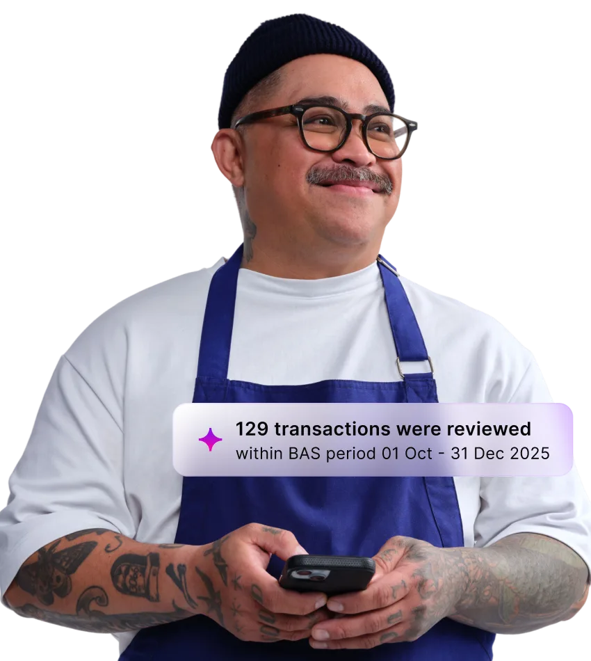 Person in blue apron and black beanie wearing glasses, holding phone with notification about 129 reviewed transactions.
