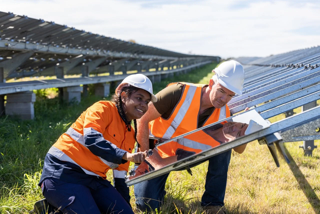 Two workers in orange safety vests and white helmets inspecting solar panels at a large solar farm installation.