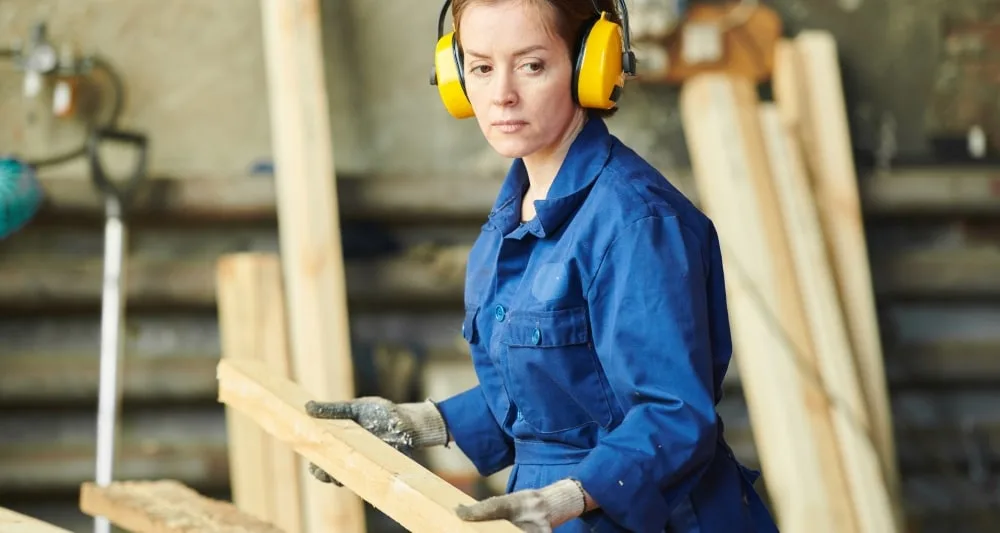A working in a workshop wearing protective earmuffs, holding a plank of wood.