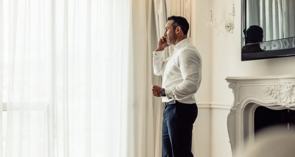 A businessperson is talking on a mobile phone whilst they are standing next to a window in a hotel room.