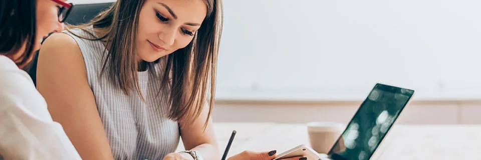 Two women collaborating at a wooden desk with laptop and smartphone, one taking notes in a bright room with natural light.