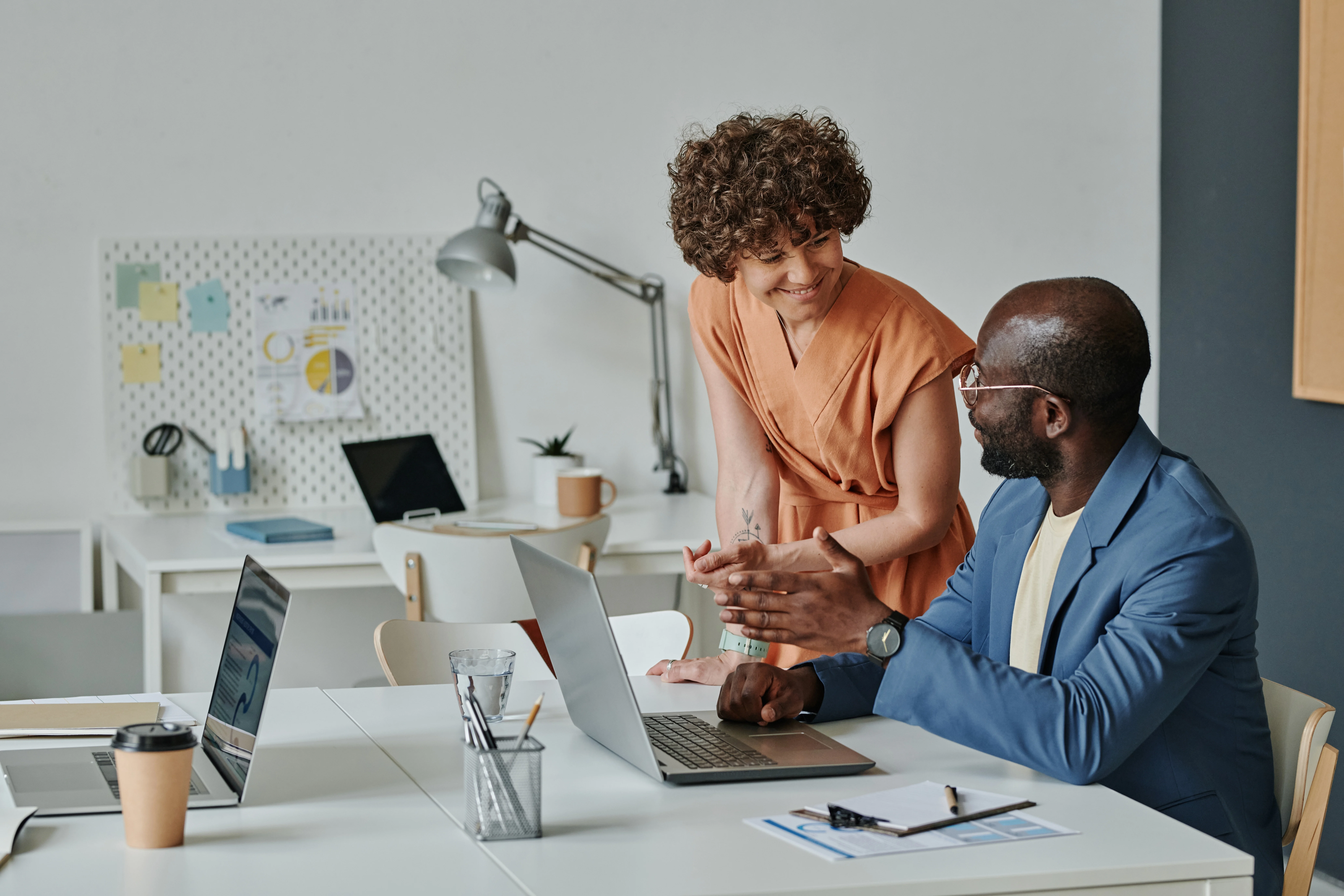 A woman with short, curly hair leans on the desk next to her business partner. The business partner, a Black man, is sitting at the desk, gesturing to his laptop. They are both smiling and confident.