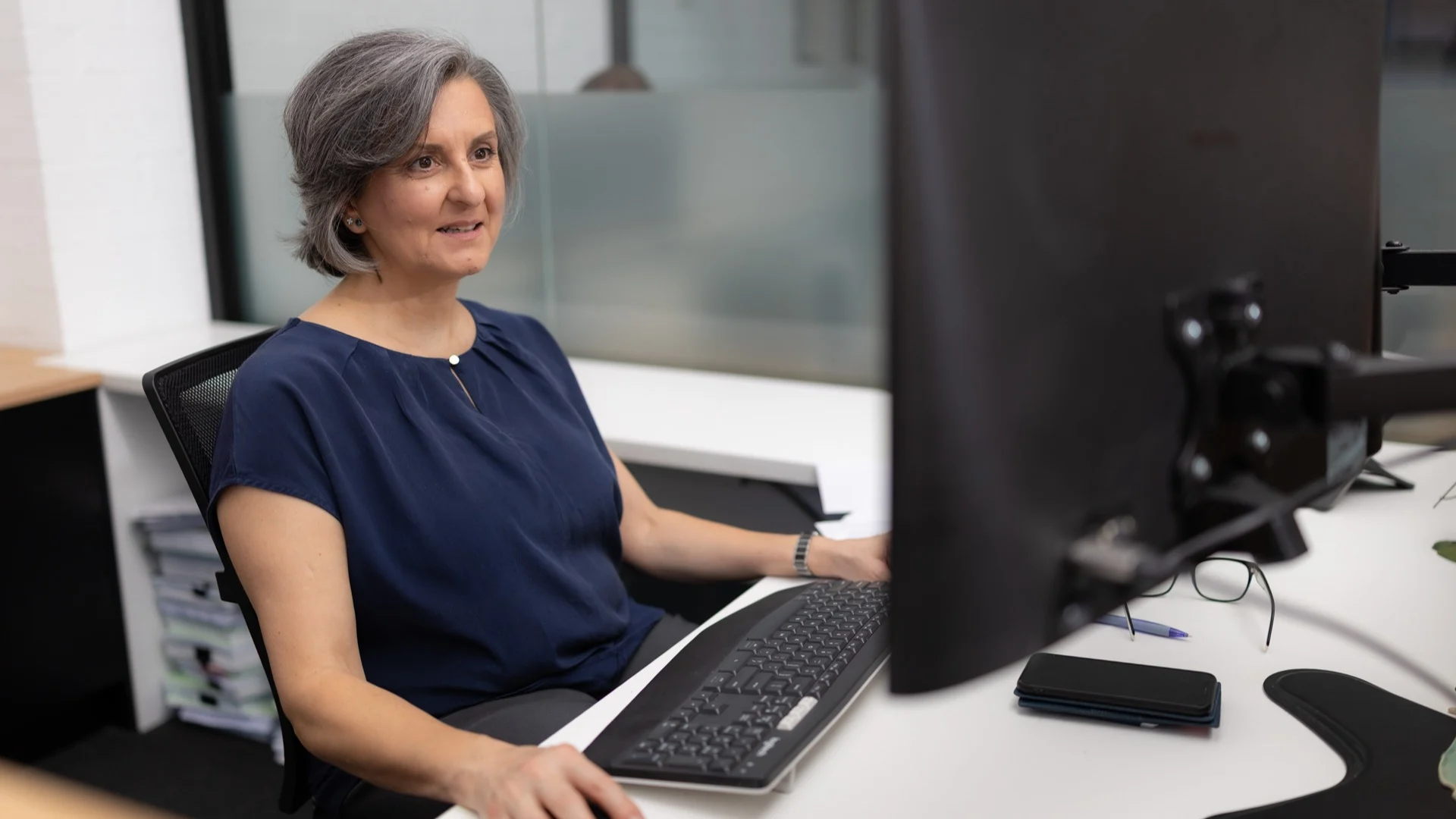 Person with gray hair in navy blue top sitting at desk with computer, keyboard and smartphone in office setting.