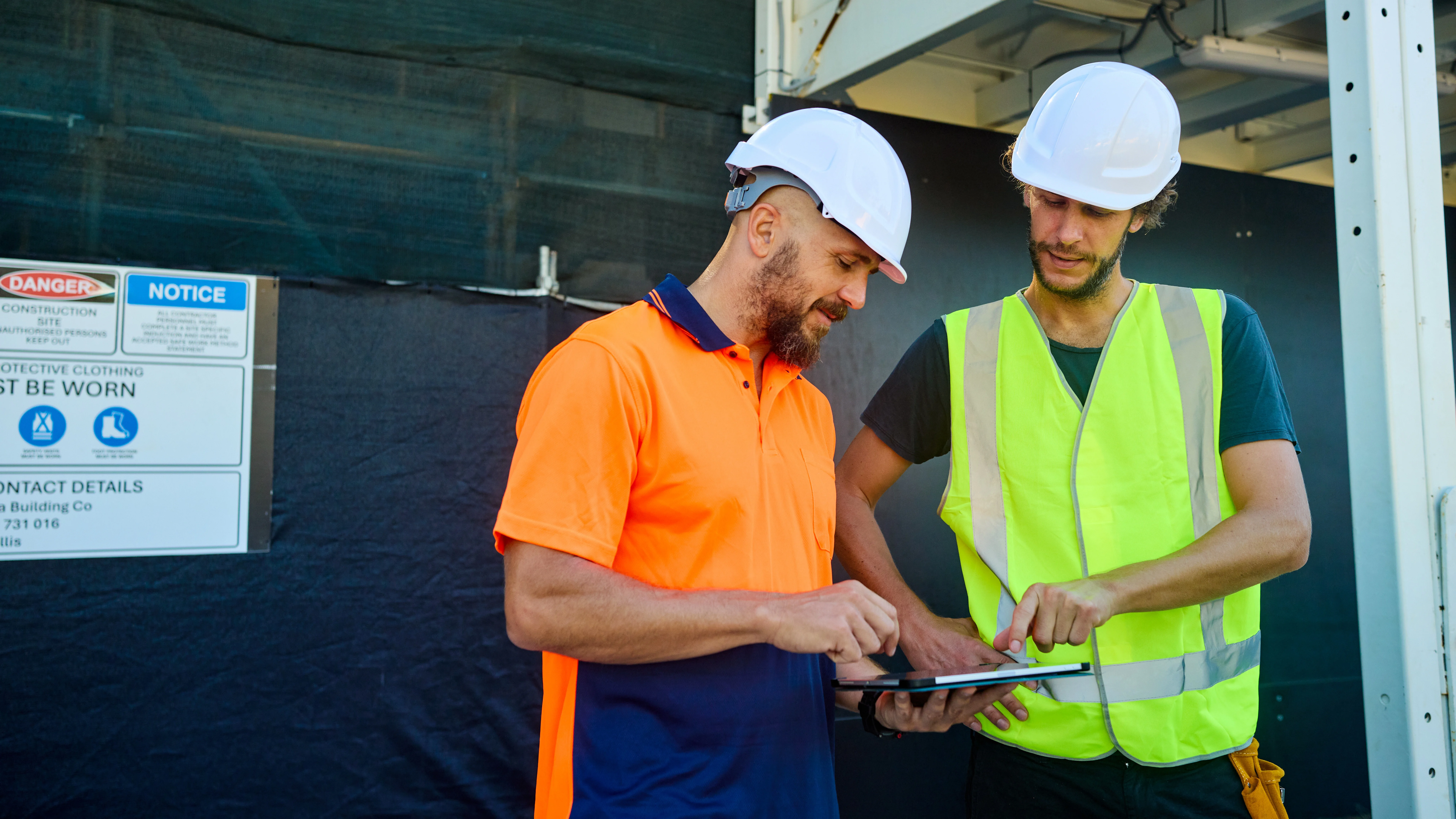 Two construction workers in hard hats and safety vests reviewing information on a tablet at a building site.