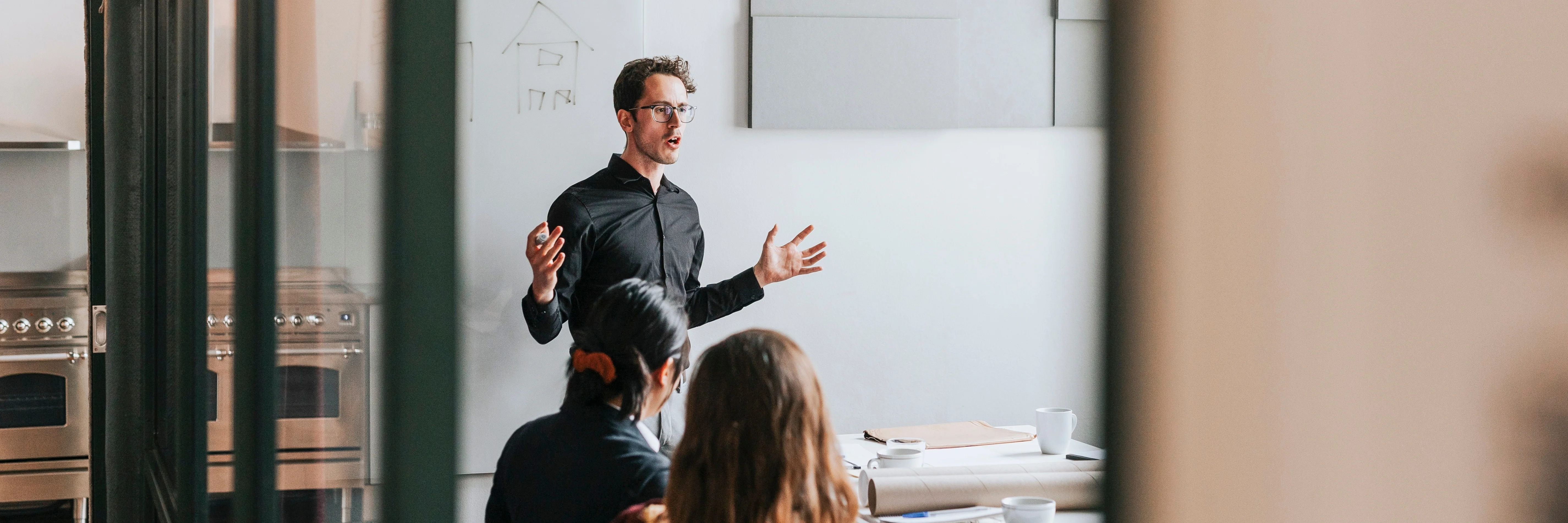 Person in black shirt presenting to small audience in modern office meeting room with whiteboard showing house sketch.