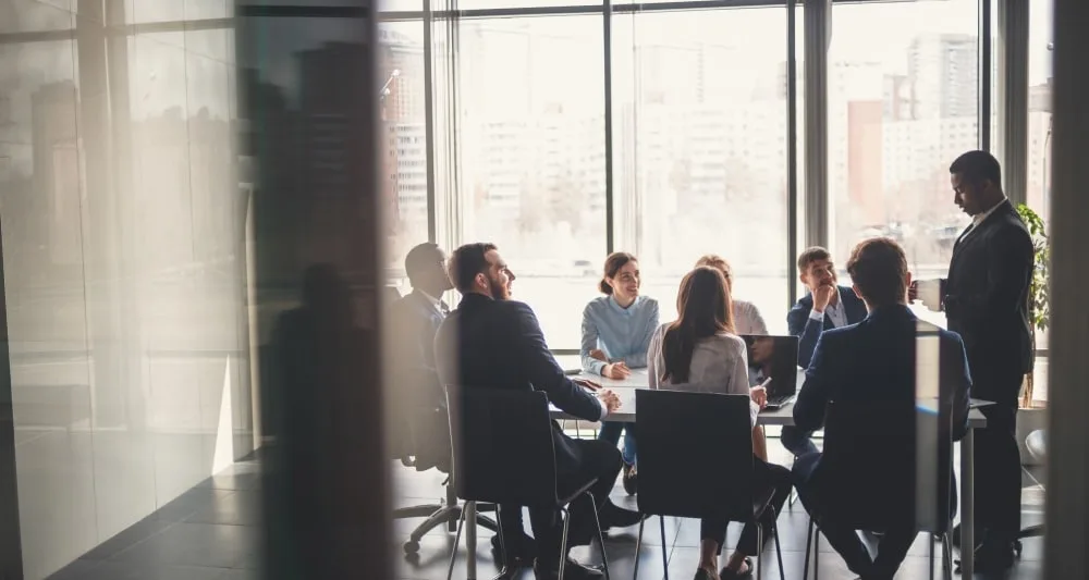 Board room table is surrounded by corporate employees watching a presenter.