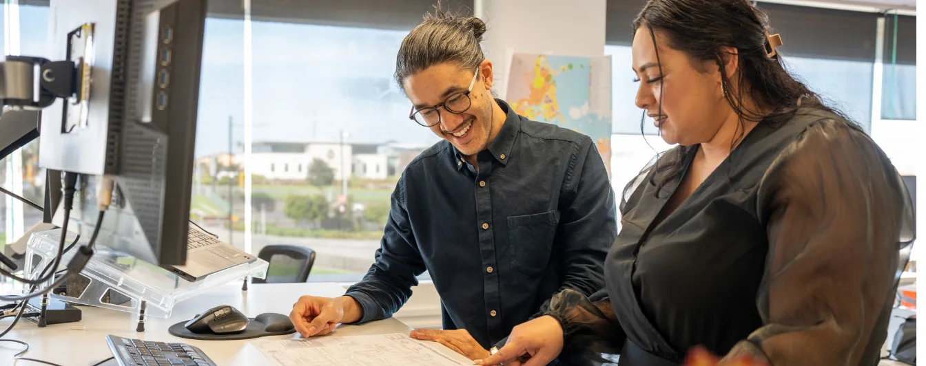 Two colleagues in dark clothing review documents together at an office desk, smiling, with monitors and a city view behind them.