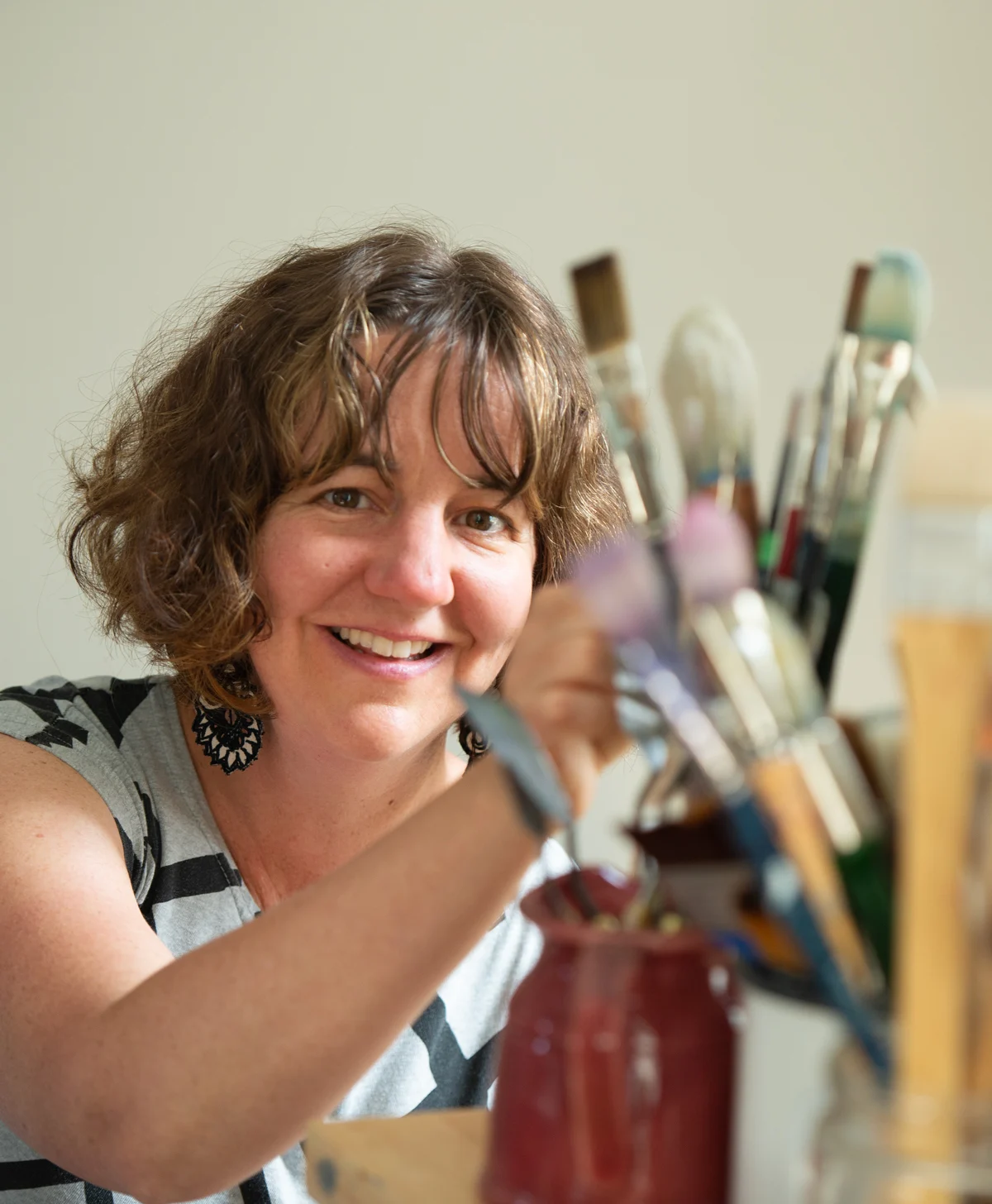A smiling woman with curly brown hair holds a paintbrush, with jars of colorful brushes in the background.