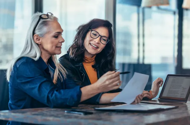 Two business people in an office sitting together at a desk. One of them holds a piece of paper and is pointing to it with a pen whilst the other person looks on smiling.