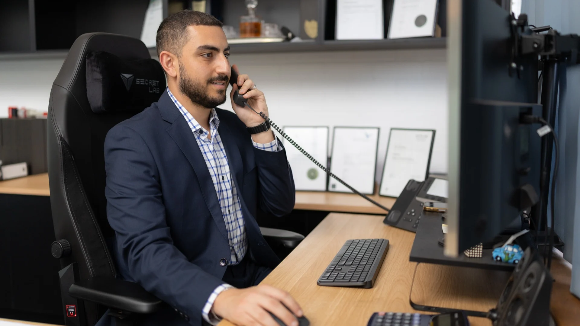 Professional in navy suit talking on desk phone in modern office with computer and framed certificates behind.