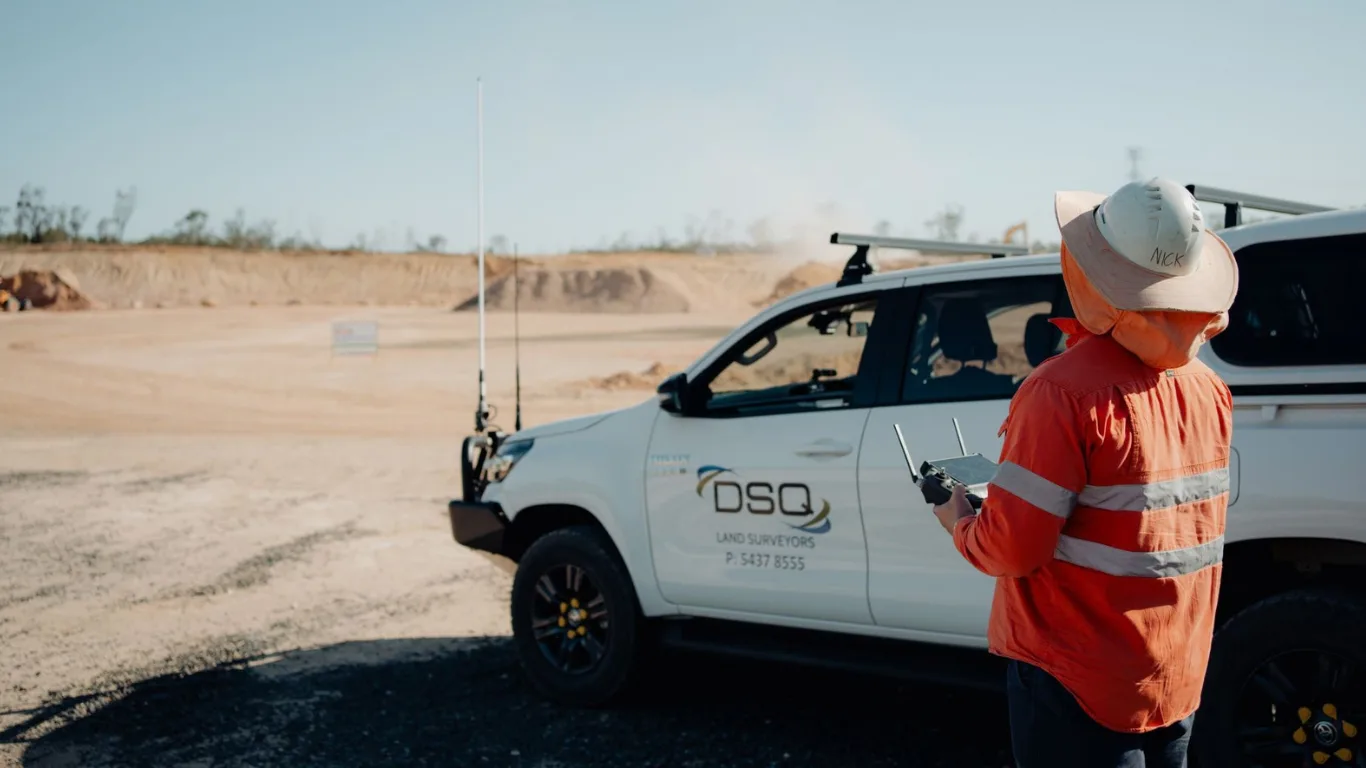 A DSQ Land Surveyors worker in orange hi-vis operates a drone controller at a mining site beside a branded white 4WD vehicle.