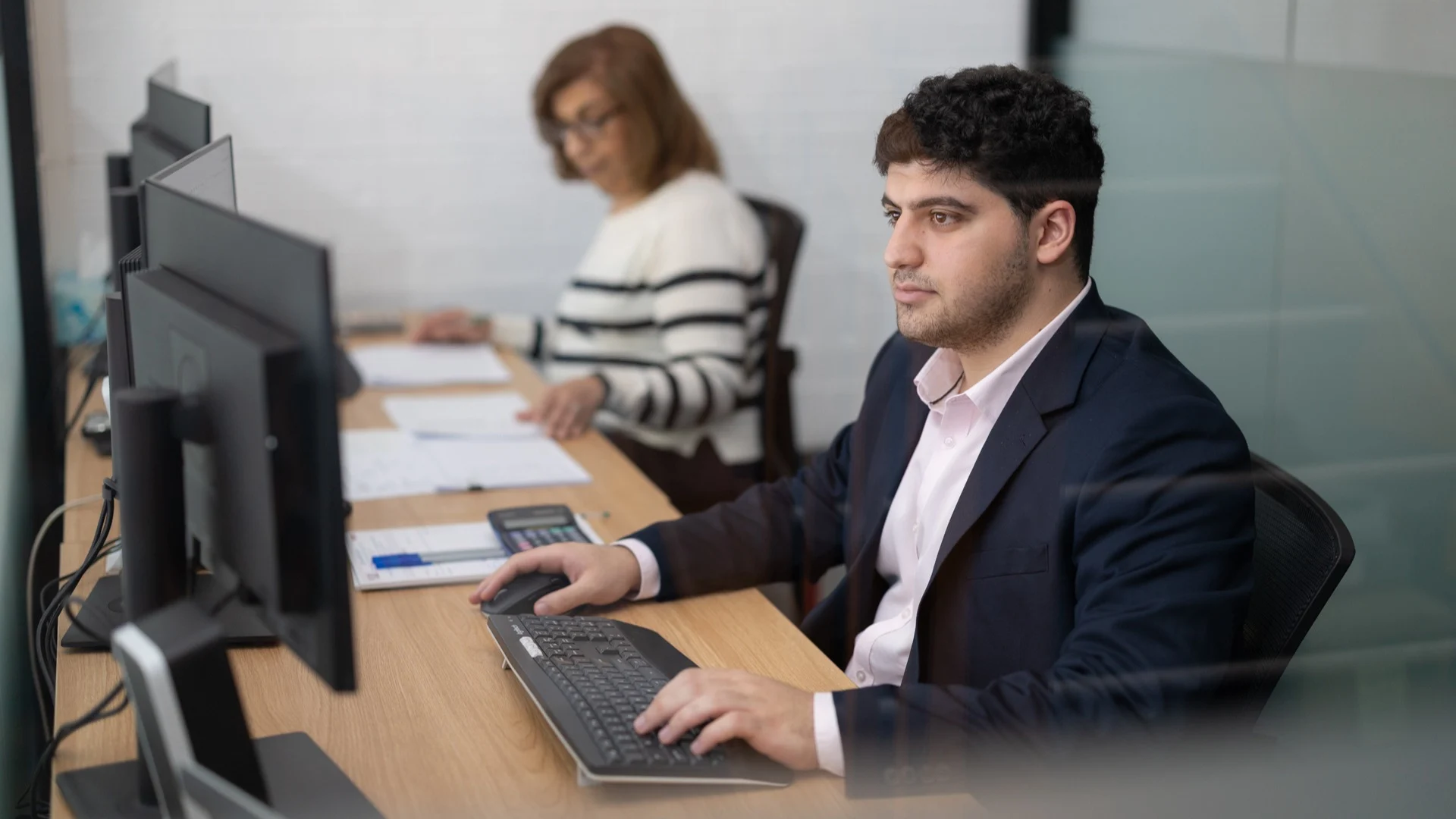 Two professionals working at computer workstations in an office, one in a navy suit and another in striped attire.